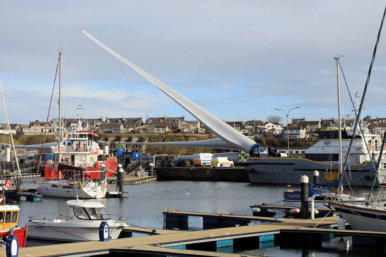 Small moored boats fill the marina foreground as a long white wind-turbine blade rests on a barge by the stone quay under an overcast sky.