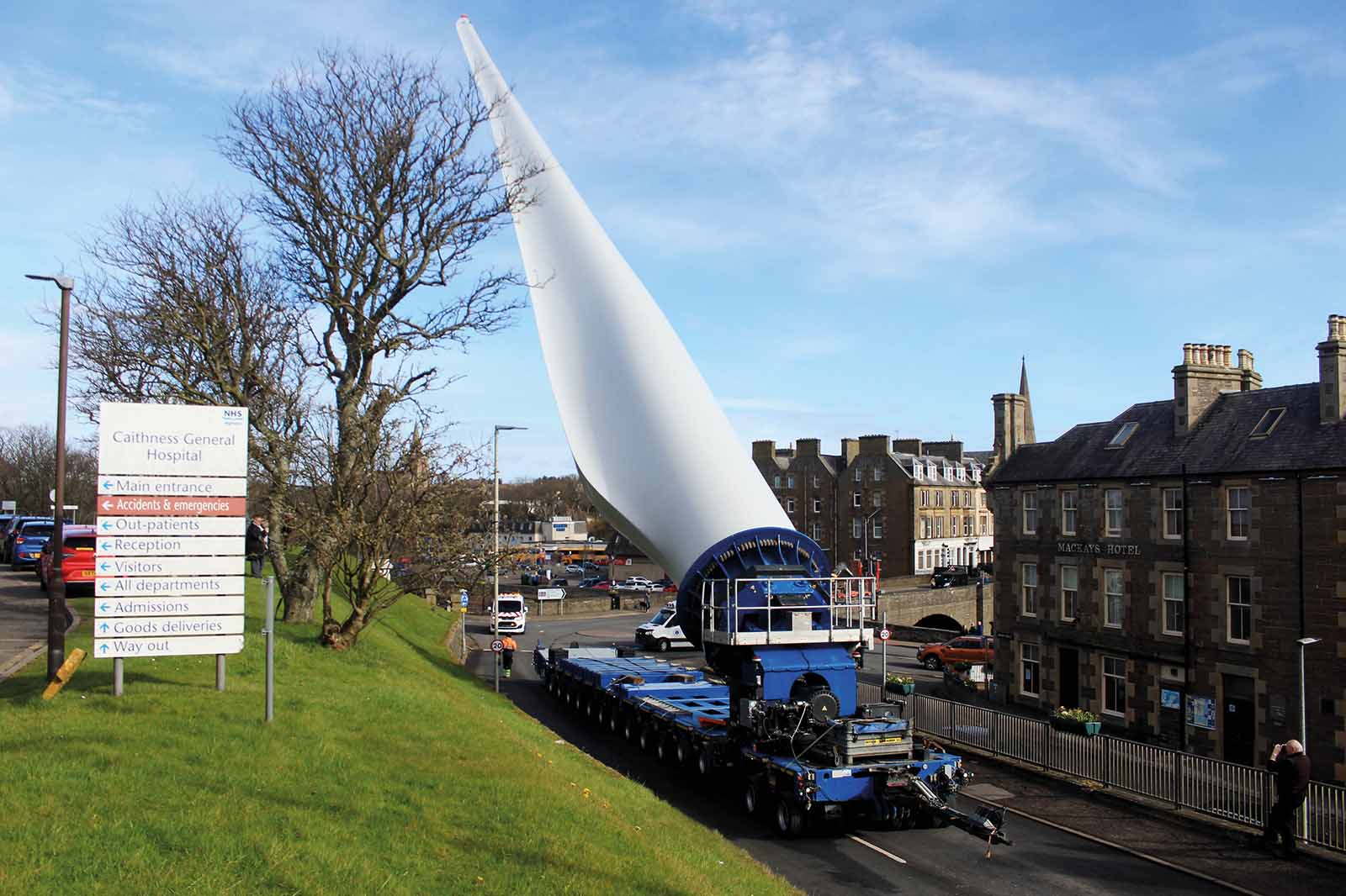 A long white wind turbine blade on a blue trailer occupies town street near stone buildings and grassy verge with a sign reading "Caithness General Hospital".
