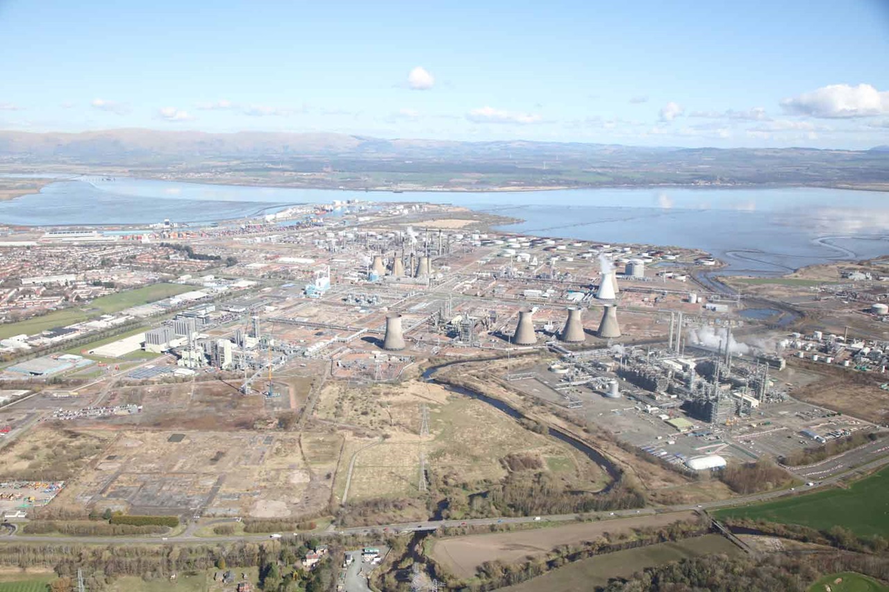 Aerial view of an industrial area featuring cooling towers, pipelines, and a river, surrounded by greenery and water.