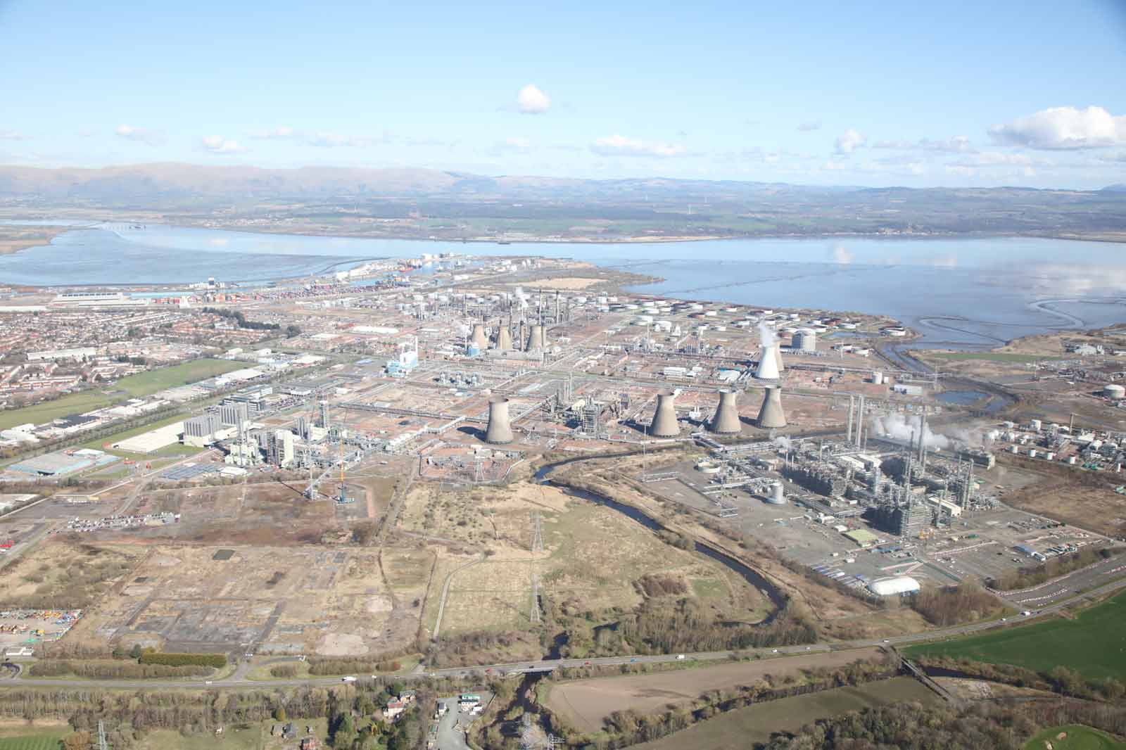 Aerial view of an industrial area featuring cooling towers, pipelines, and a river, surrounded by greenery and water.