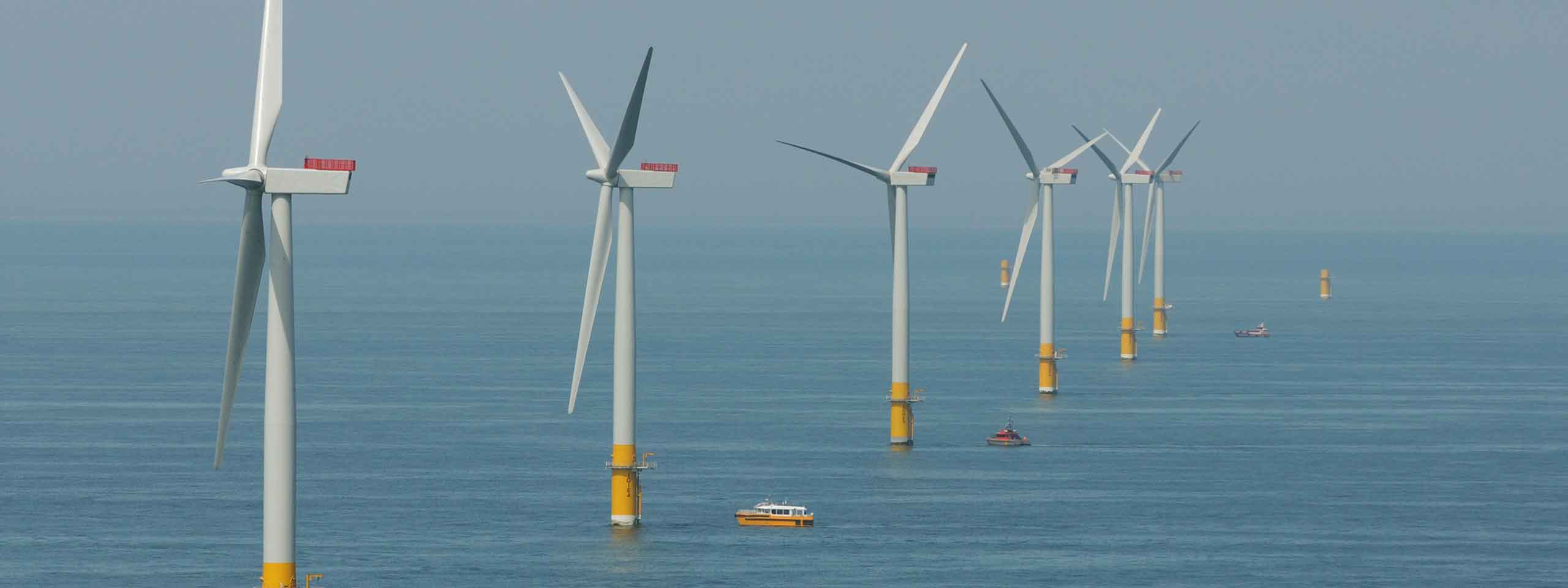 Offshore wind turbines in the sea with a small boat nearby. The sky is clear and the water is calm.