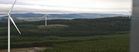 A view of wind turbines in a green forested landscape with rolling hills in the background under a cloudy sky.