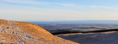 A scenic view of rolling hills and a coastal landscape with wind turbines in the distance under a clear blue sky.