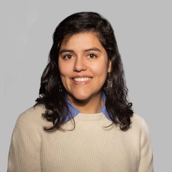 A woman with dark, curly hair wearing a cream sweater over a blue collared shirt, standing against a plain grey background. Portrait of  Ghislaine Morales Arroe.