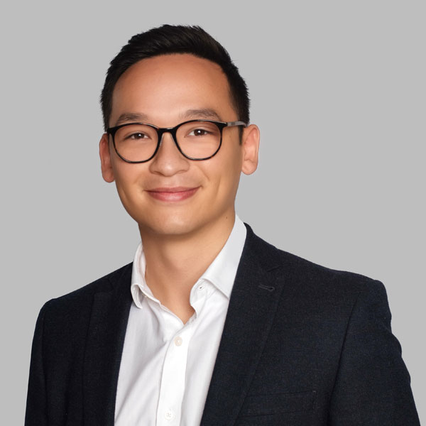 A man in a dark blazer and white shirt with round black glasses is posing against a grey background.