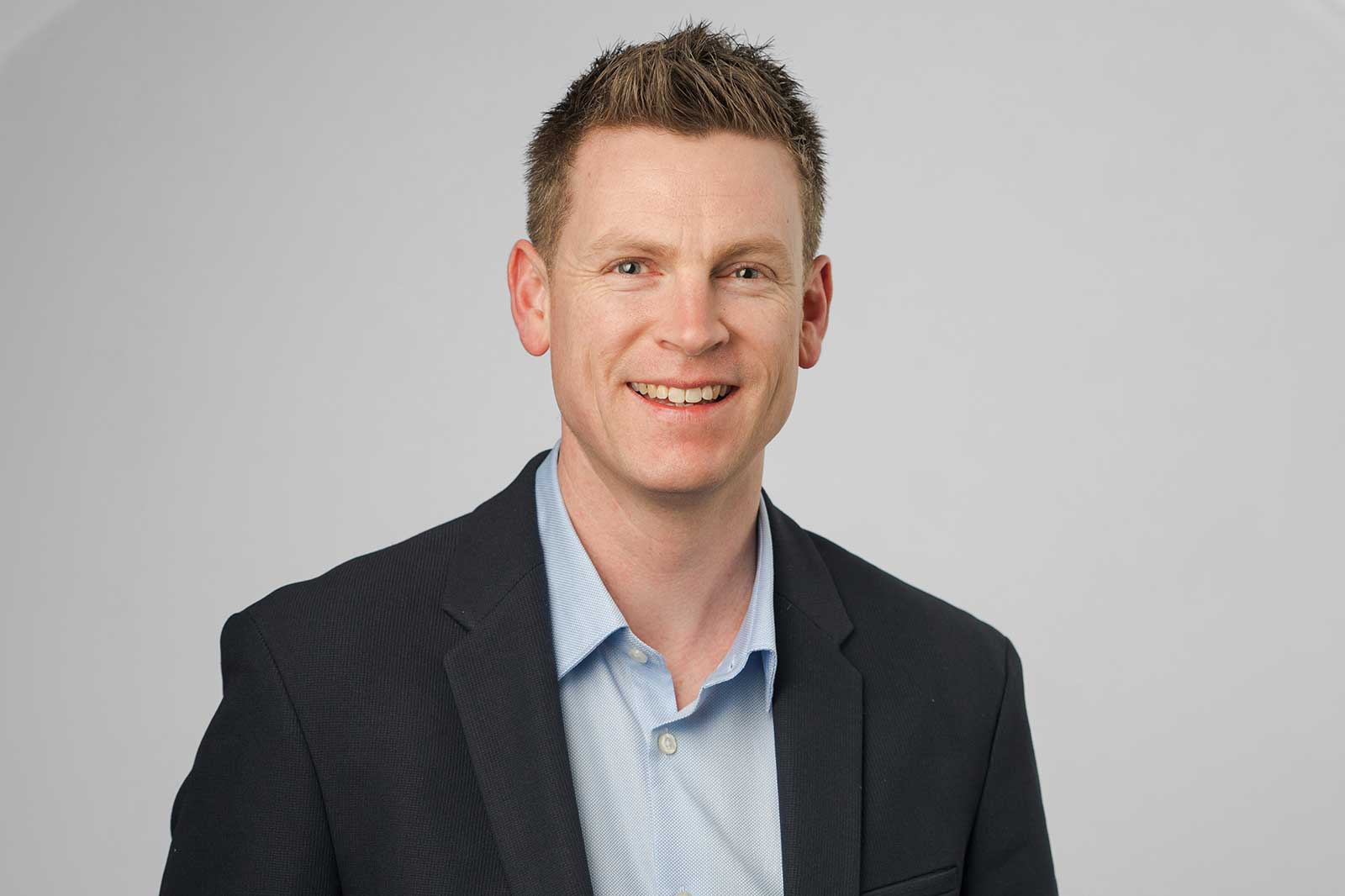 A man in a black blazer and light blue shirt poses against a plain background.