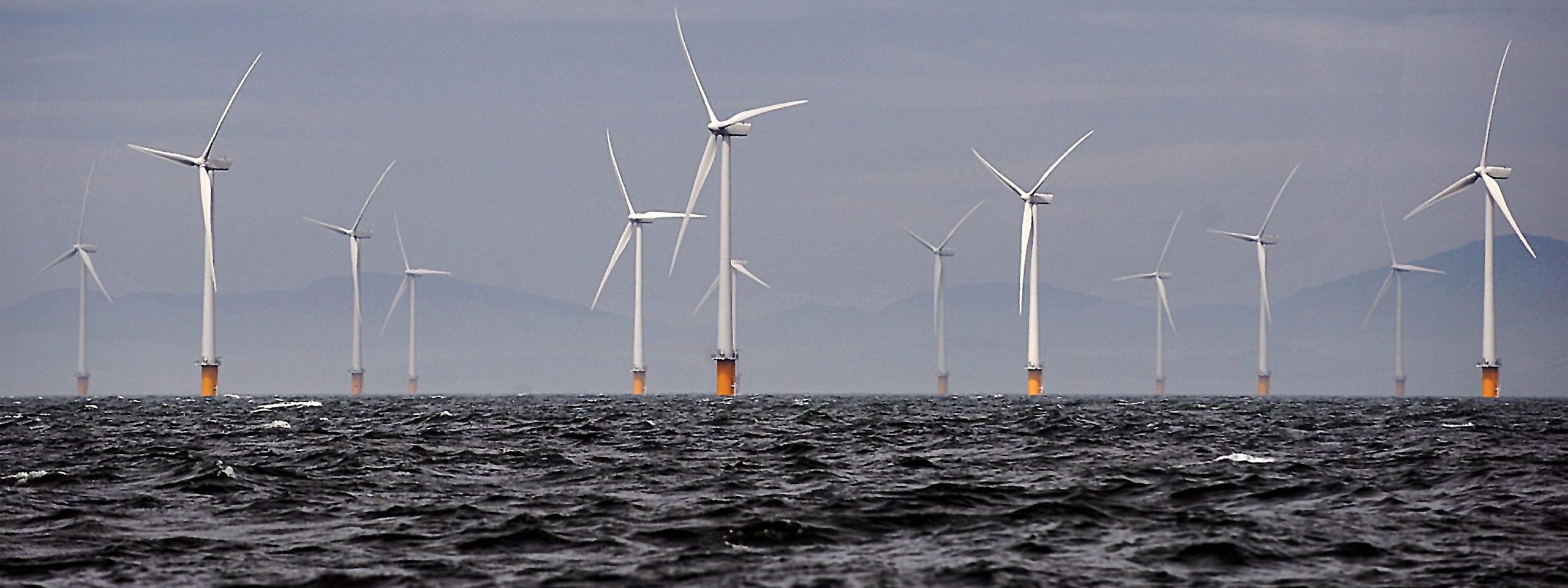 Offshore wind turbines stand tall above the waves, set against a cloudy sky with distant hills.