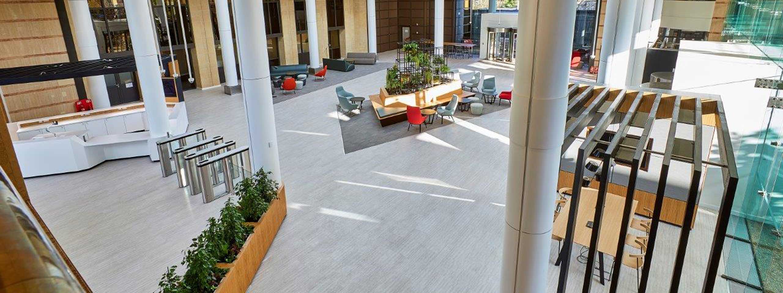 A spacious modern lobby with seating areas, plants, and large windows, featuring white pillars and sleek furniture.