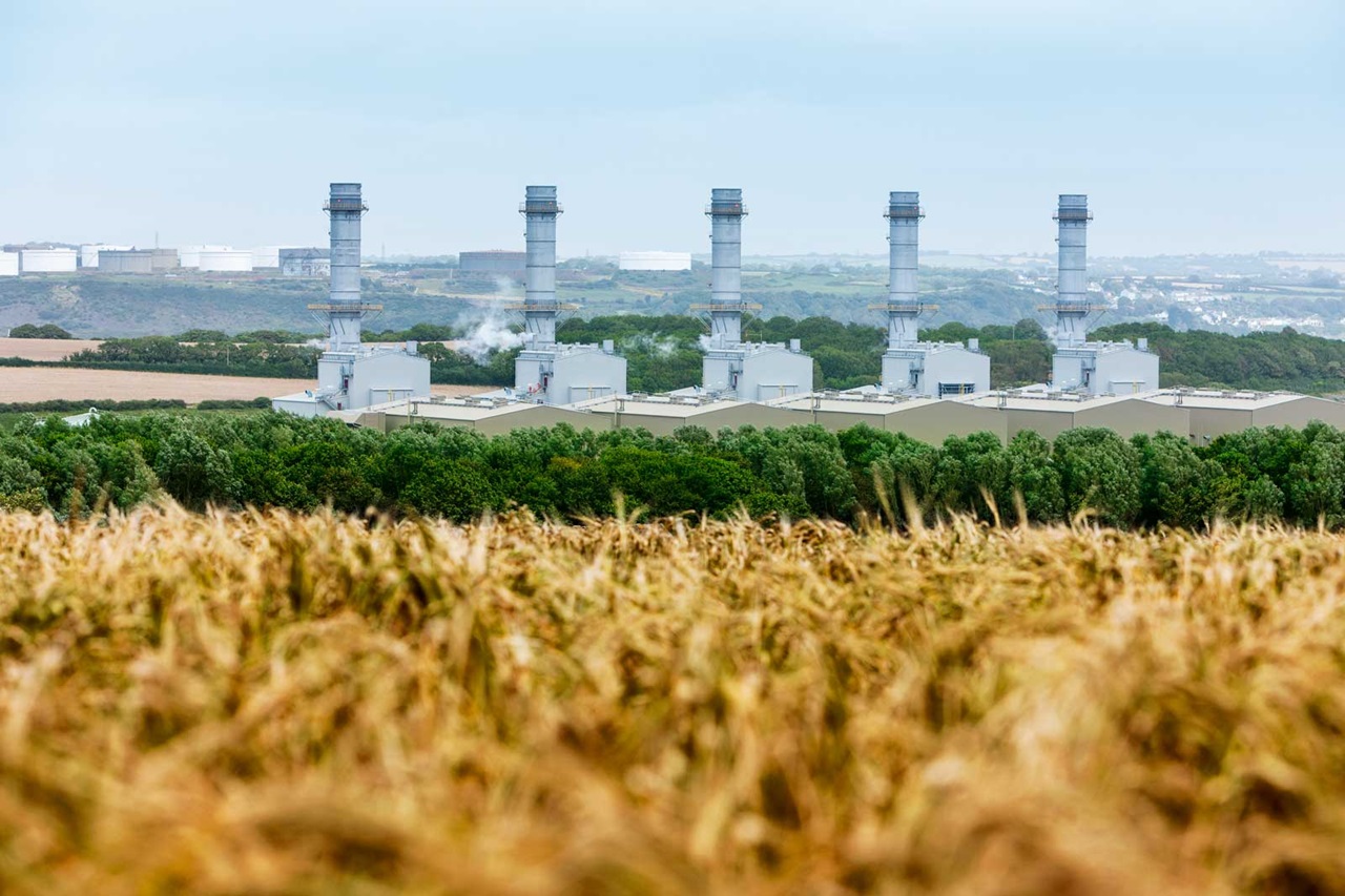 A power plant with four tall smokestacks is set against a green landscape, surrounded by fields and trees under a cloudy sky.