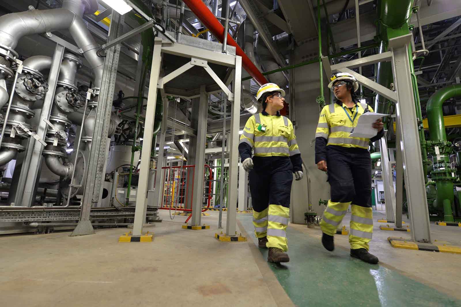 Two workers in safety gear walk through an industrial facility, surrounded by large pipes and machinery.
