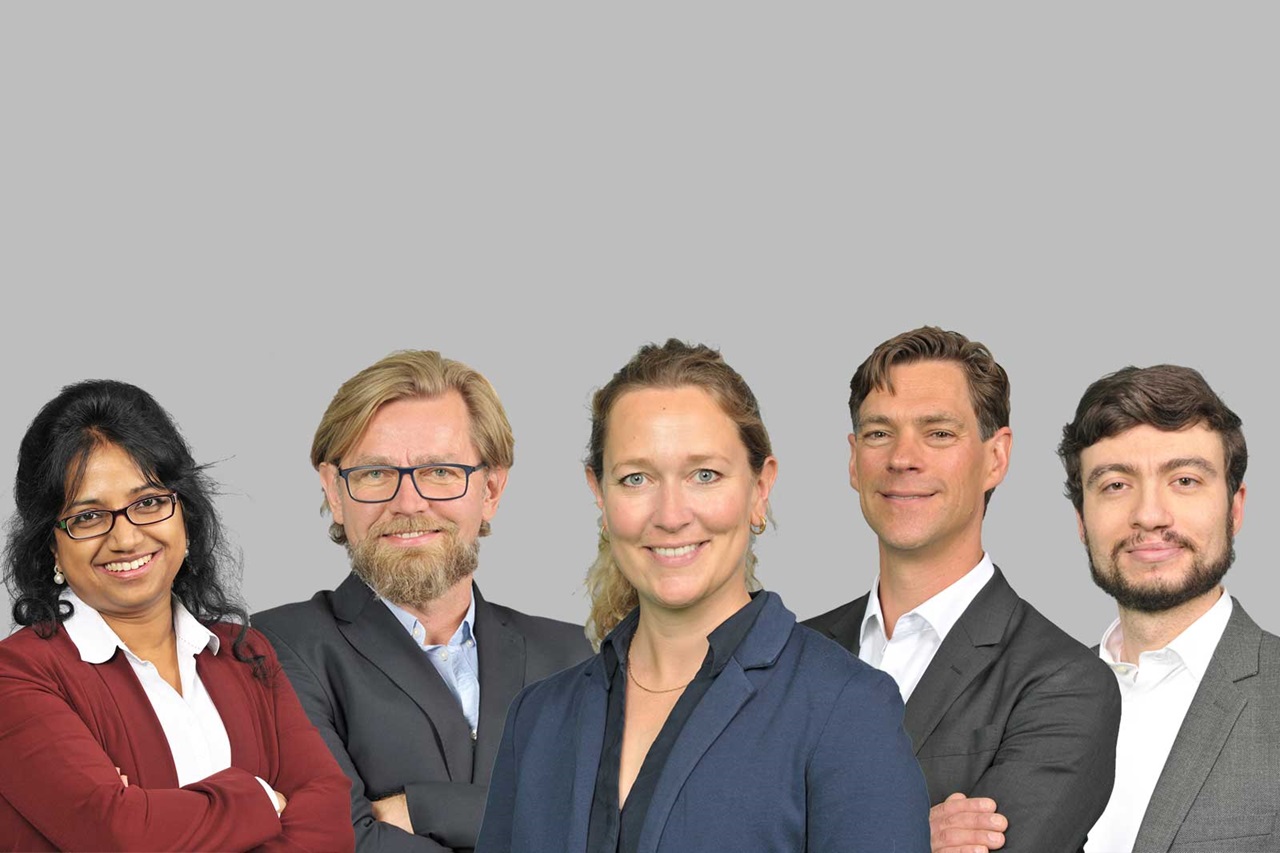 A group of five individuals in formal clothing against a grey background.