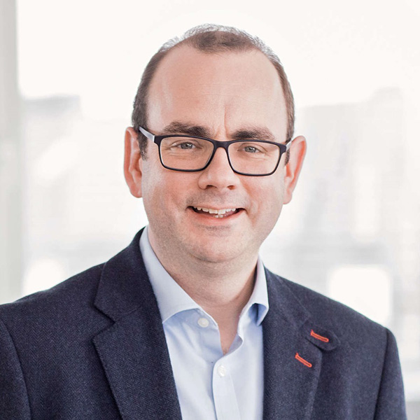A man in a dark blue blazer with a red accent stands in a bright room with a blurred background, portrait of Tom Glover.
