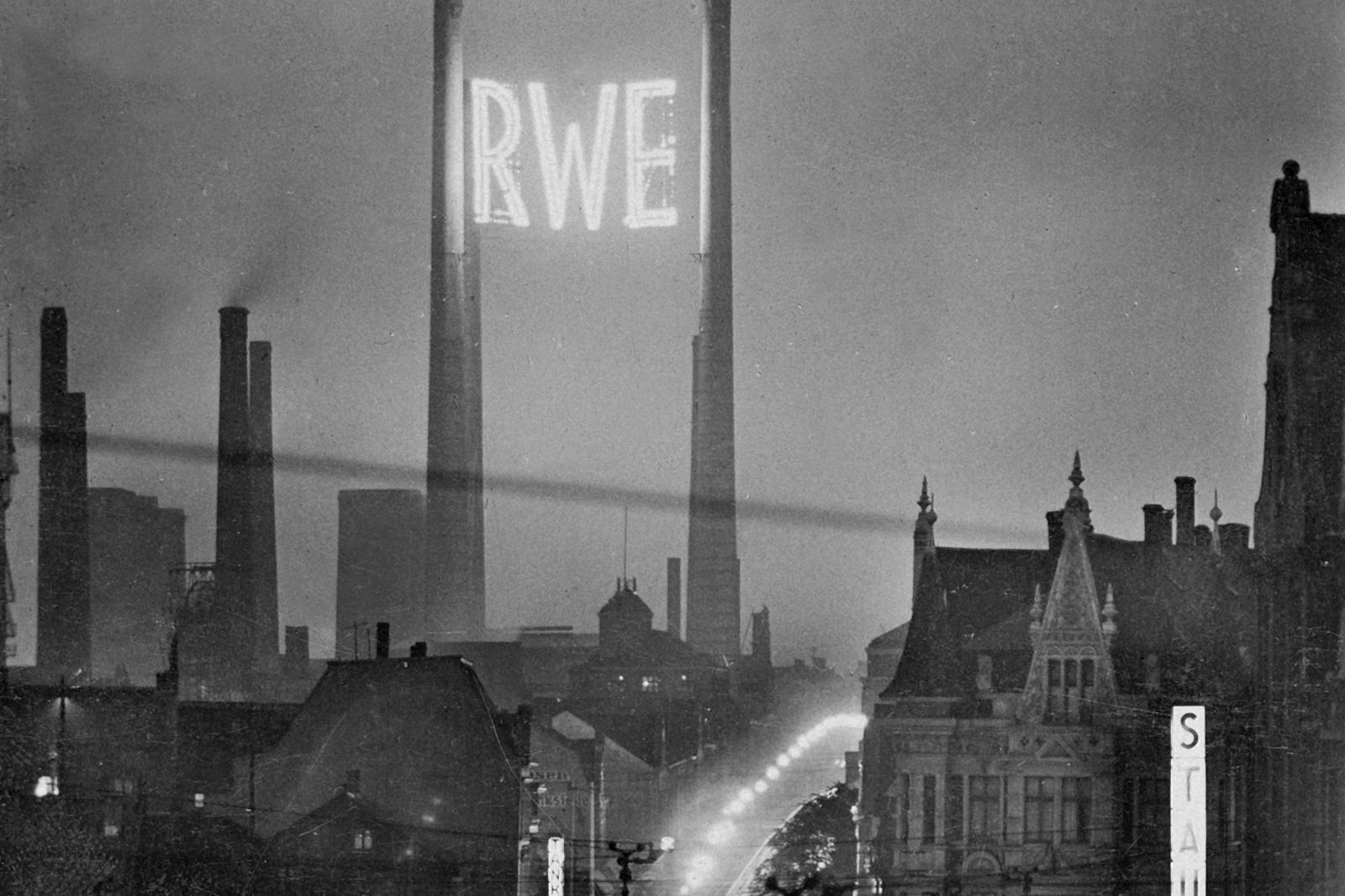 A historical black-and-white photograph of a city featuring tall chimneys and the illuminated text 'RWE'.