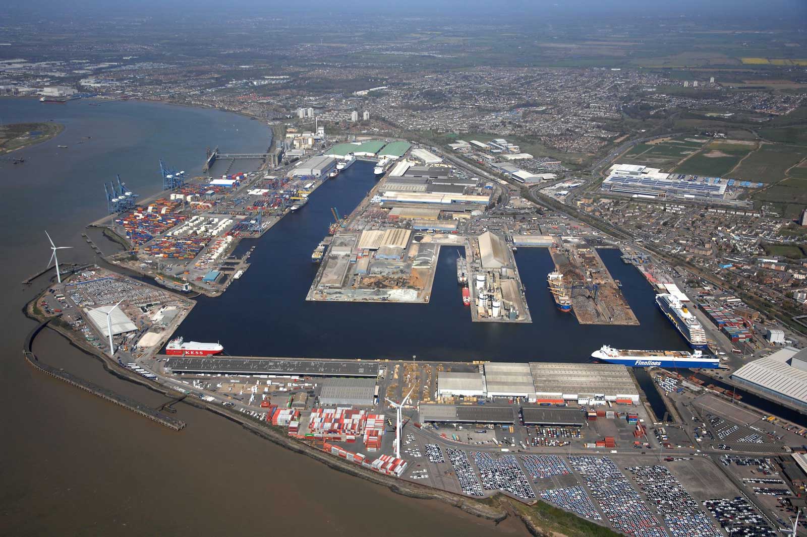 Aerial view of a busy port with shipping containers, docked vessels, wind turbines, and industrial buildings along a river.