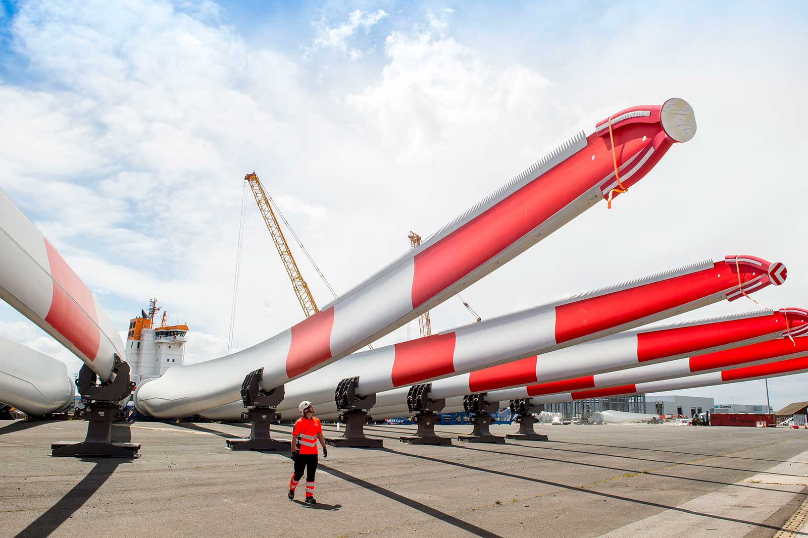 A worker in a helmet and high-visibility clothing walks between large wind turbine blades at a dock.
