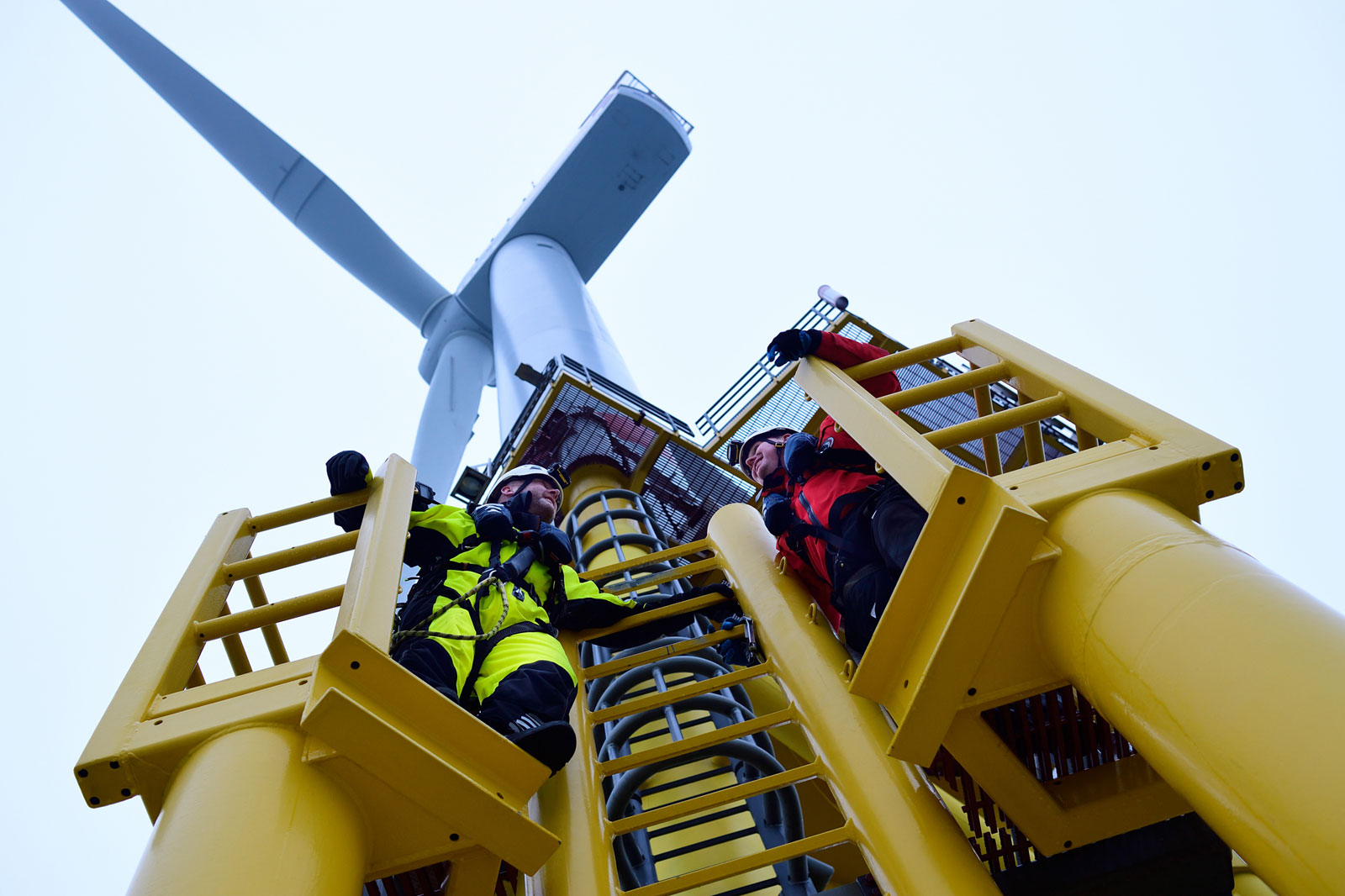 Two workers in safety gear climb a yellow ladder on a wind turbine platform, with the turbine blades above them.