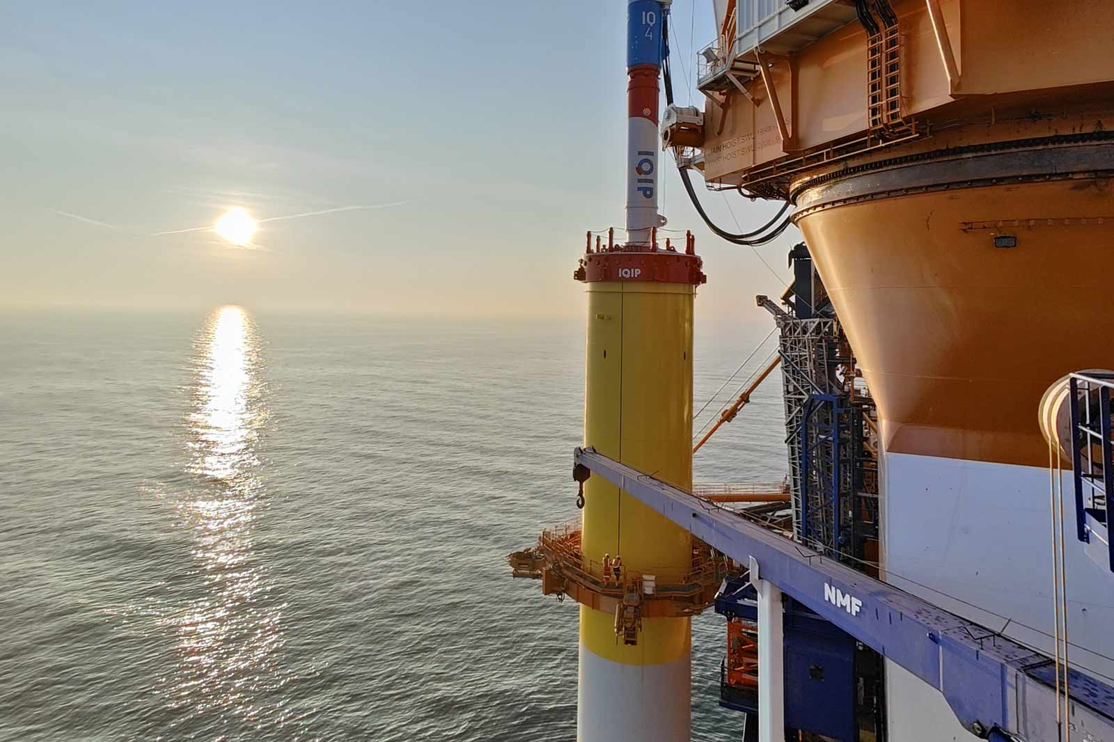 An offshore structure with yellow and white columns, silhouetted against a sunset over calm waters.