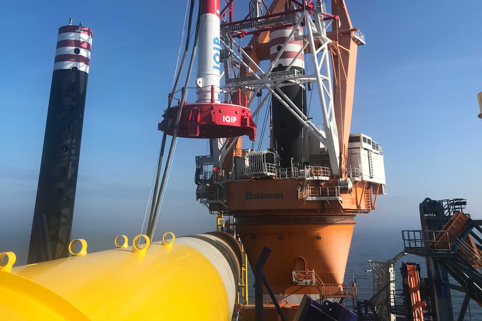 A view of an offshore construction platform with large equipment and a bright yellow pipe under a clear blue sky.