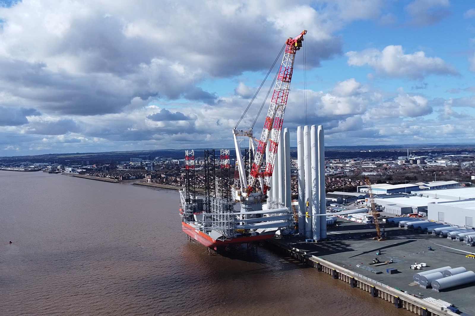A large crane vessel is docked at a riverside, surrounded by an urban landscape with clouds in the sky.