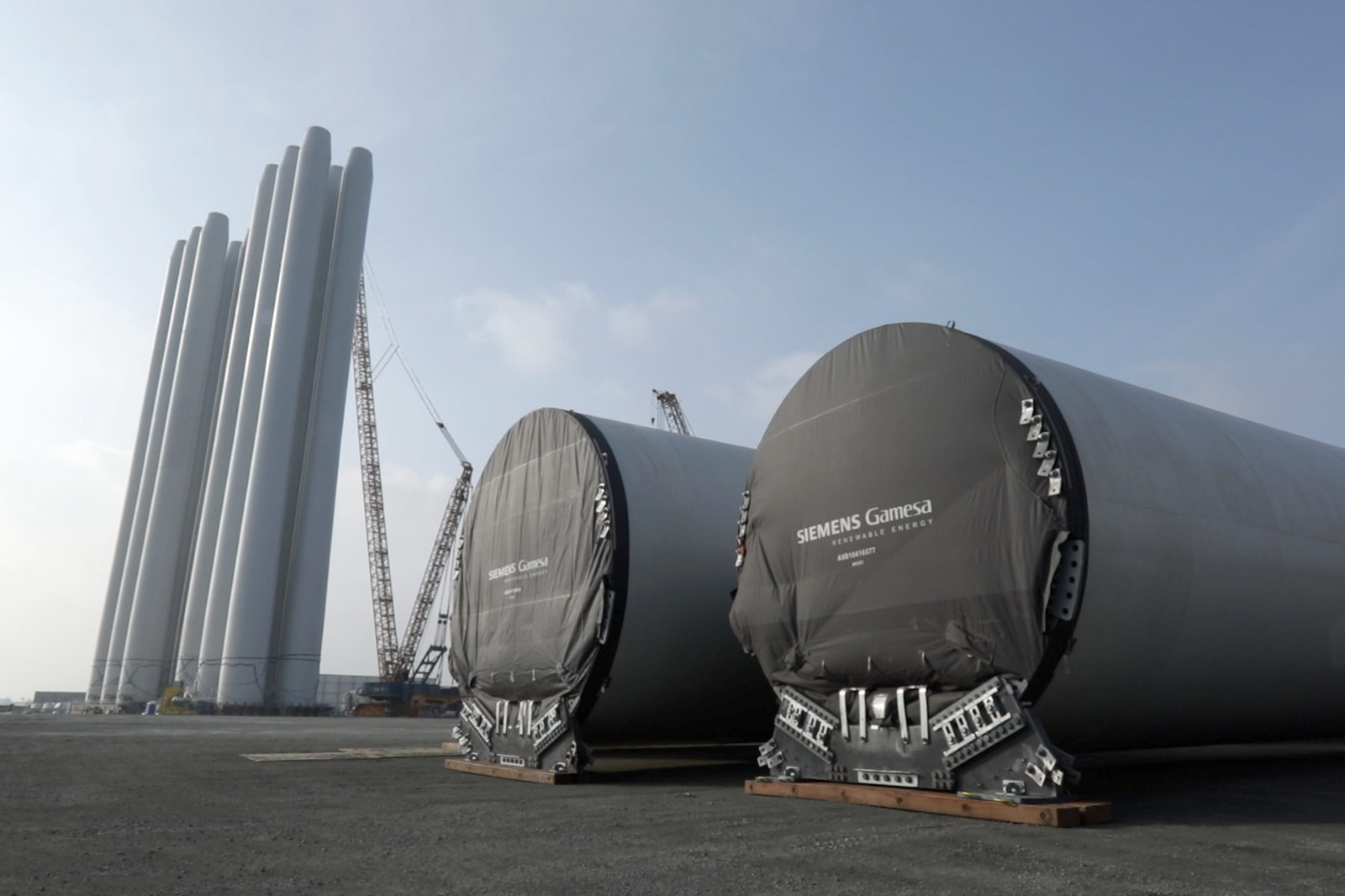Two large wind turbine blades are wrapped in black covers, standing side by side on a construction site, with tall columns in the background.