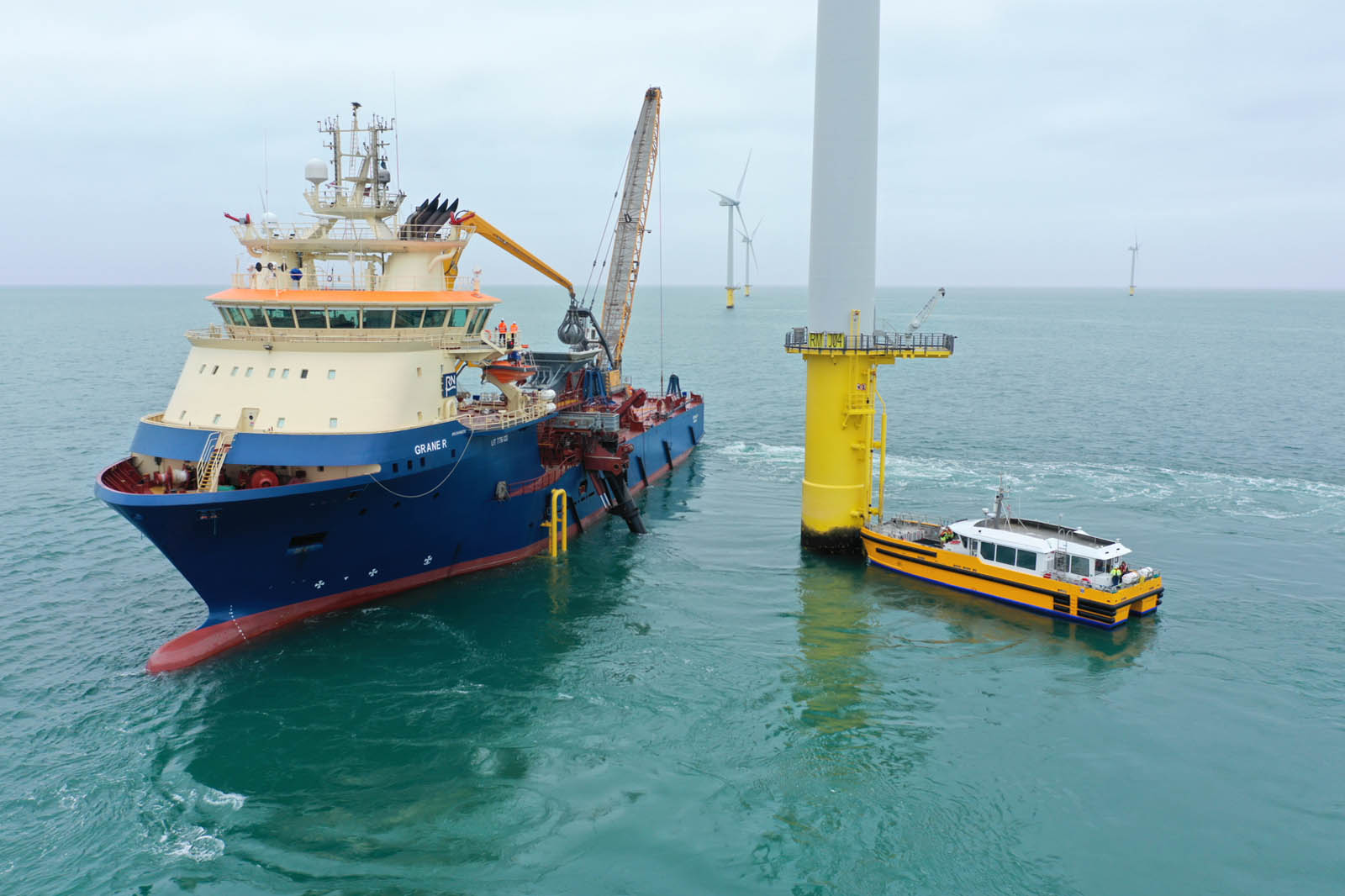A large support vessel, Grane R, is docked near a yellow offshore wind turbine, with a smaller boat alongside.