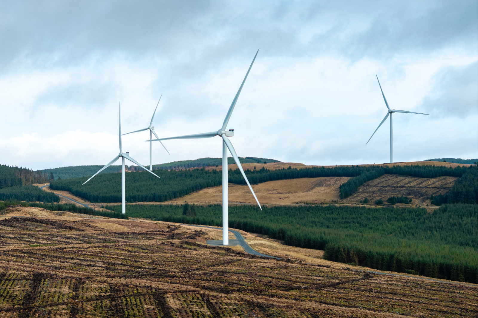 A landscape with several wind turbines on a hillside, surrounded by green forests and cloudy skies.