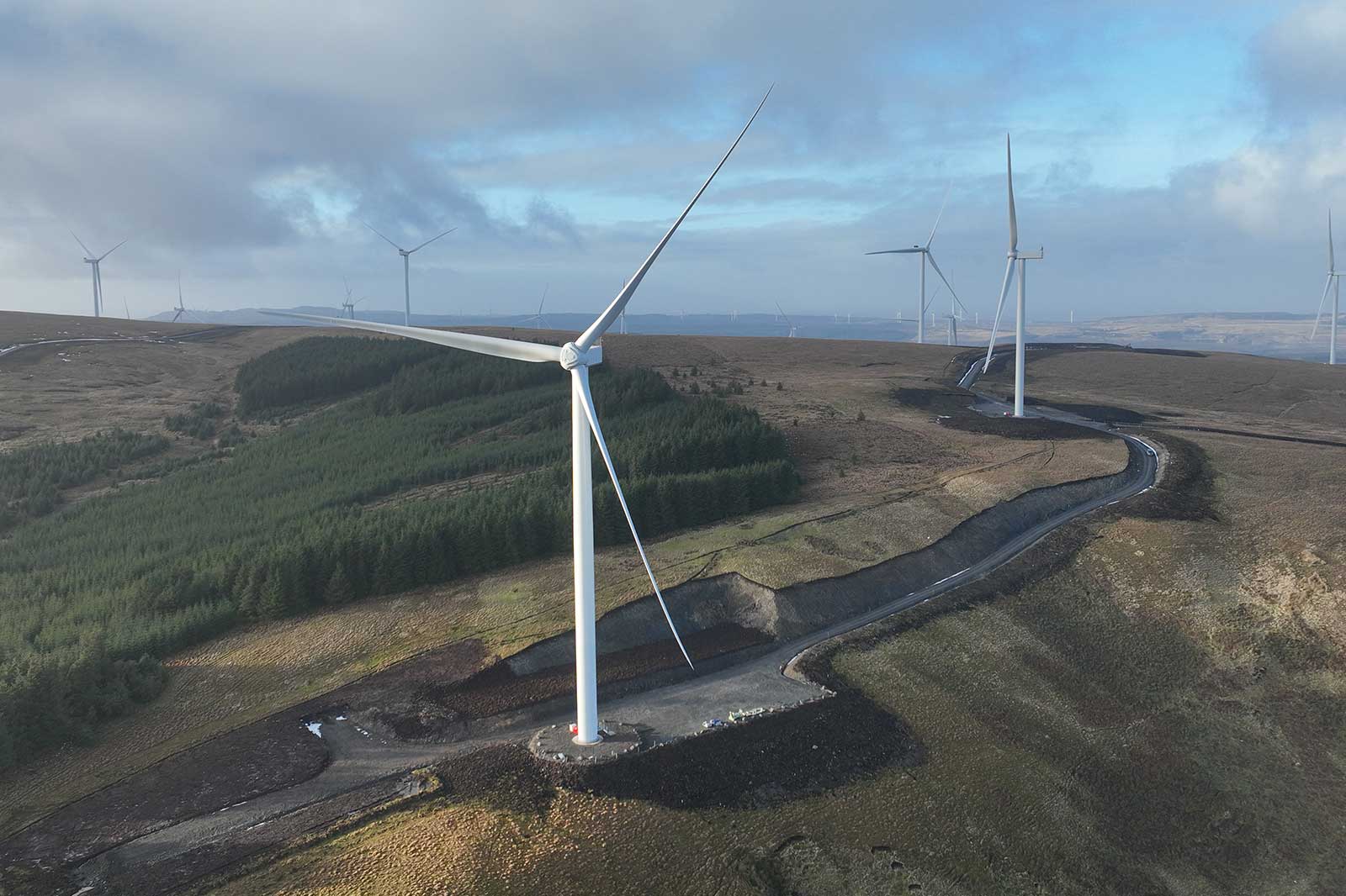 A white wind turbine on a concrete pad dominates brown and green moorland, with a winding gray road and turbines beneath a cloudy blue sky.