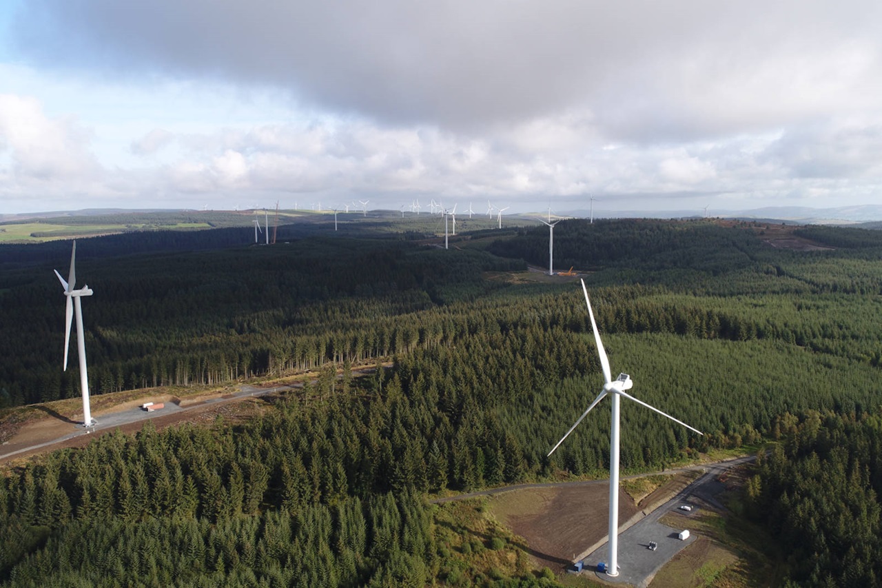 Aerial view of numerous wind turbines situated amidst a dense forest landscape under a cloudy sky.