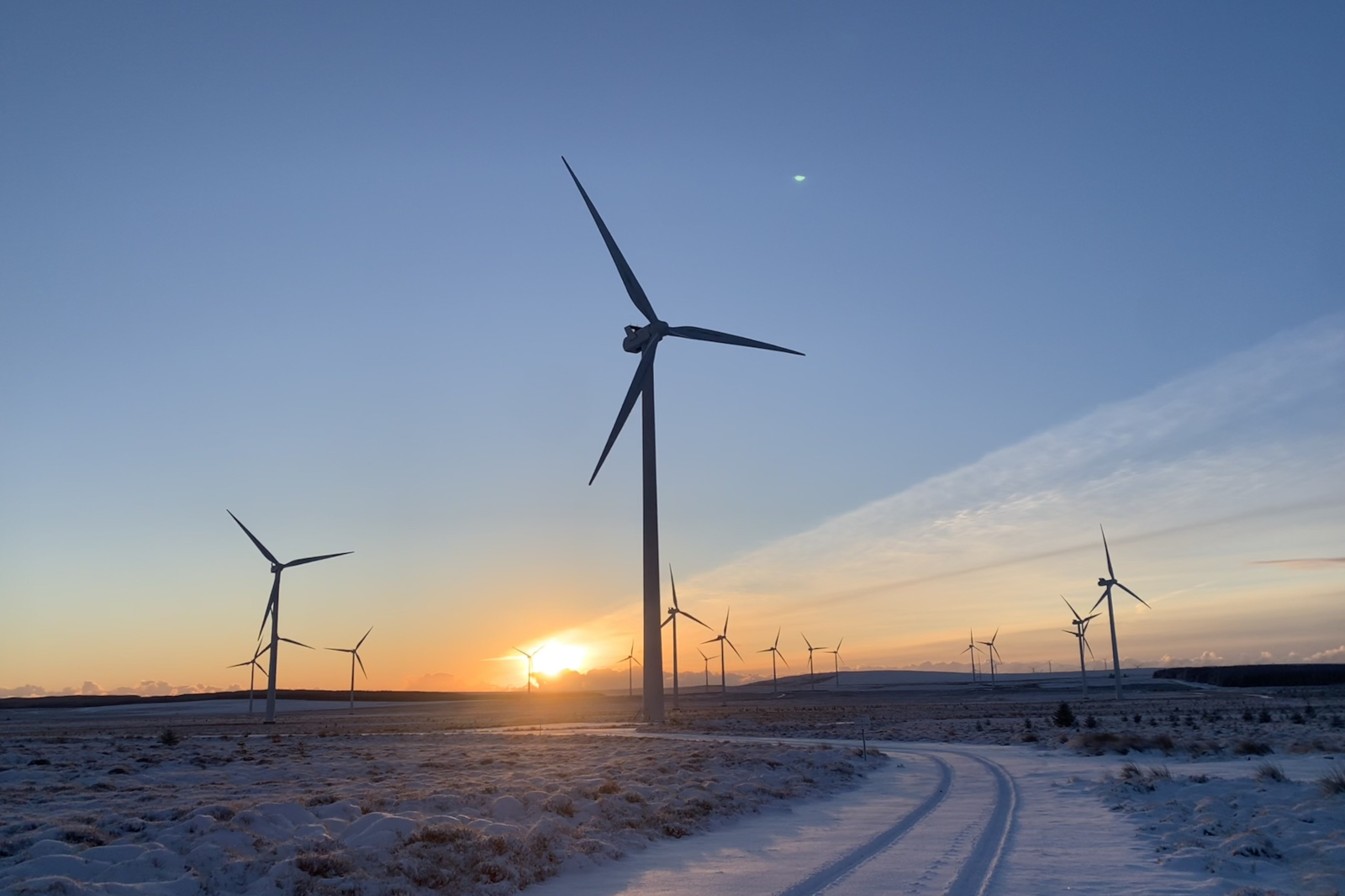 A winter landscape featuring wind turbines silhouetted against a glowing sunset.