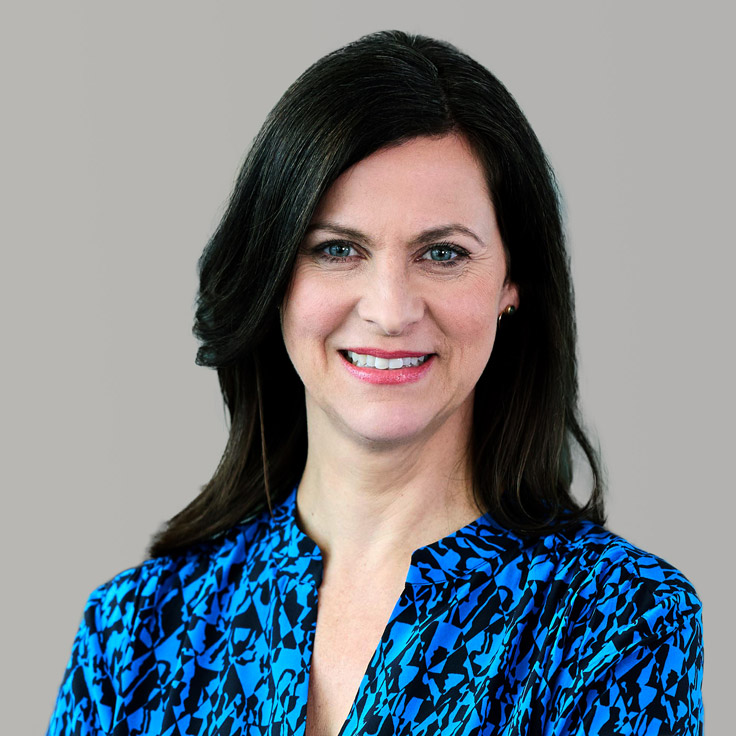 A person with long dark hair wears a blue patterned blouse. The background is grey. Portrait von Birgit Hiller.