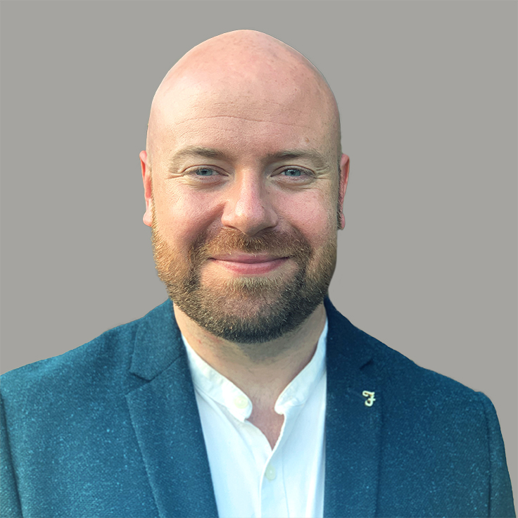 A person in a dark blue blazer and white shirt stands against a grey background. Portrait of Kyle Raistrick.