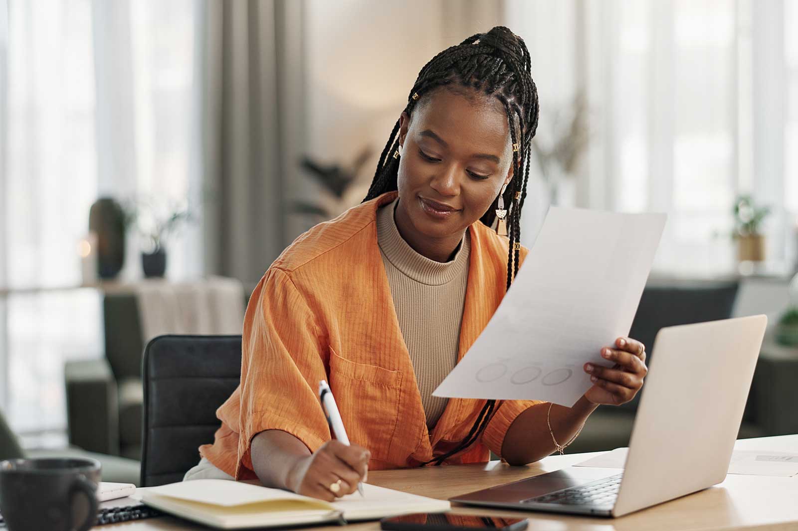 A person with braided hair sits at a desk, holding a document and writing on their laptop.