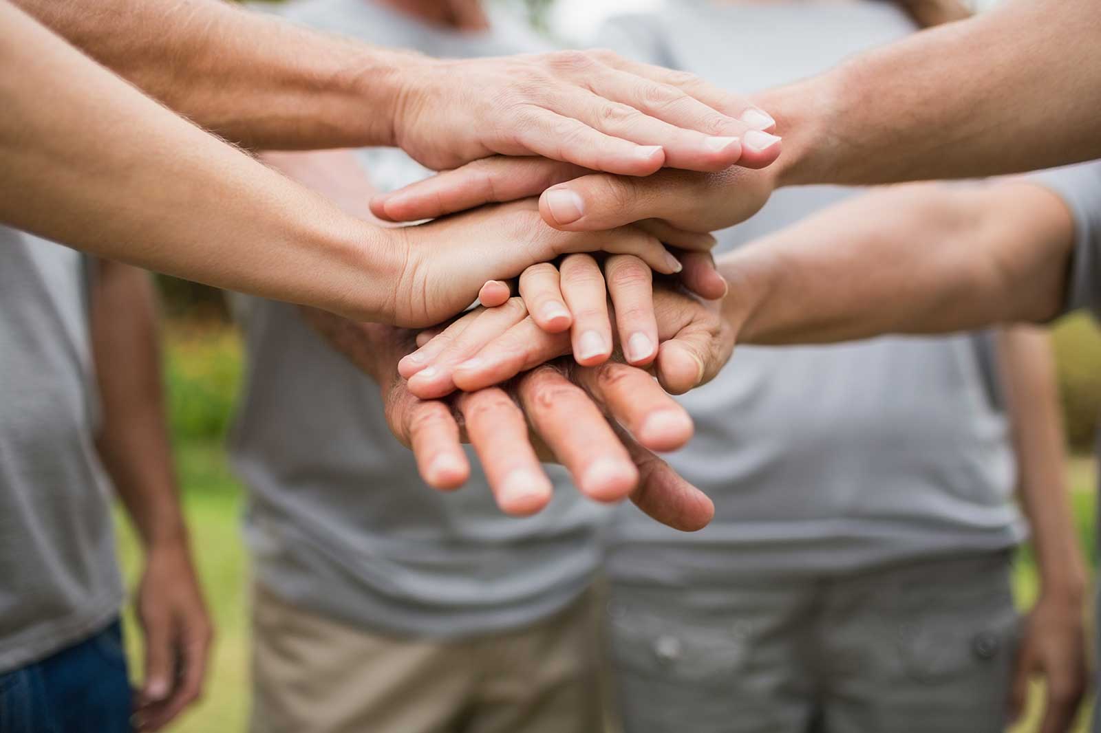 Several adults stack their hands in a team huddle, wearing grey T-shirts and standing outdoors.