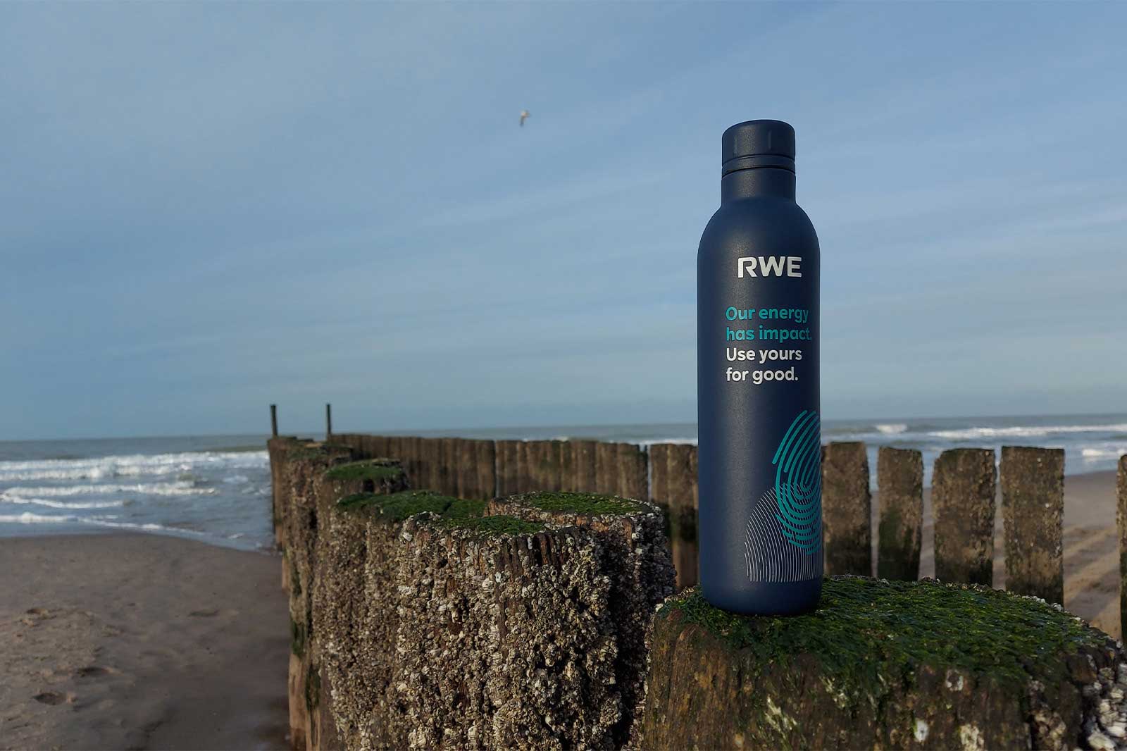 A reusable water bottle with the RWE logo stands on a weathered wooden post on a beach, with ocean waves in the background.
