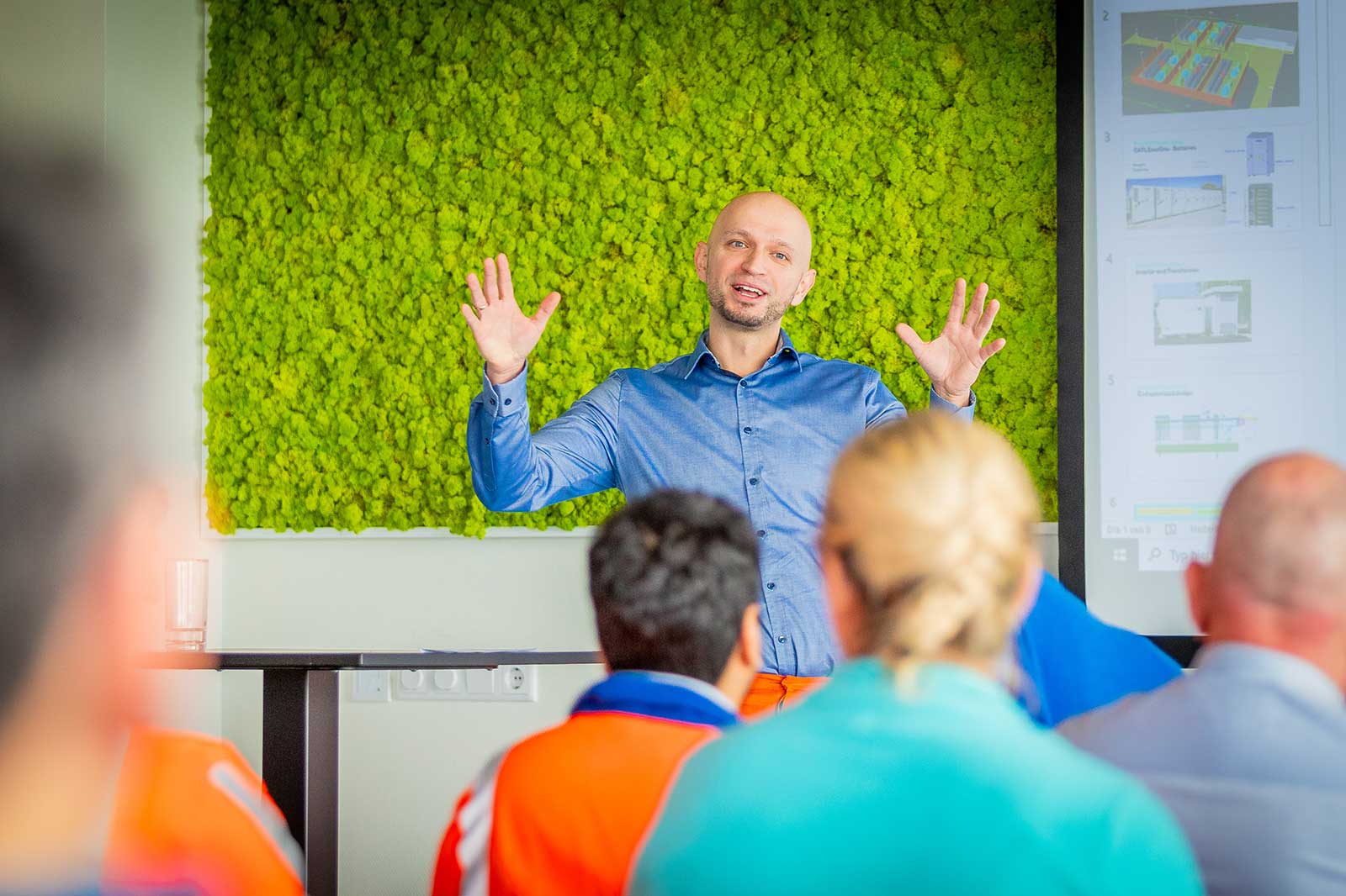 A speaker gestures enthusiastically during a presentation in front of a green moss wall, with an audience engaged in discussion.