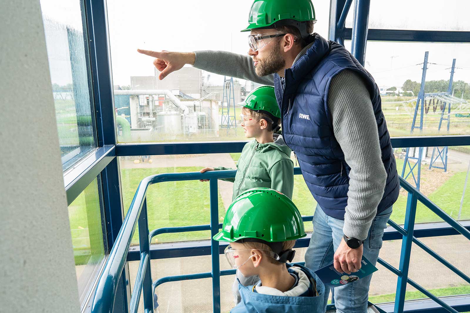 A man points out the view to two children, all wearing green safety helmets, in a building with large windows.