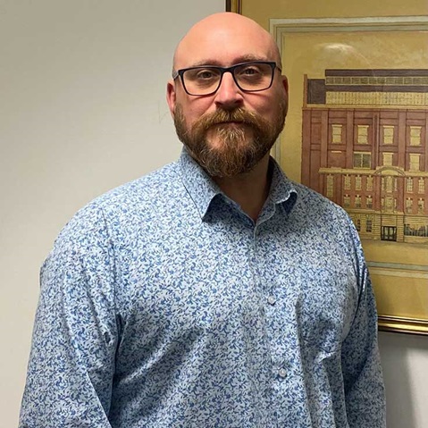 A person in a blue floral shirt stands in front of a framed picture of a building.