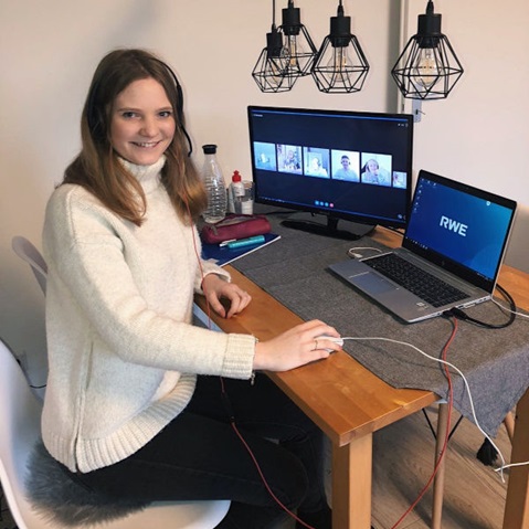 A person sits at a table with two computers. In the background, there are lamps and water bottles visible.