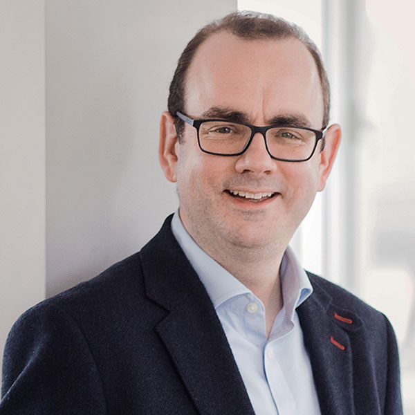 A man in a navy blazer and light blue shirt stands against a light wall, showcasing a professional appearance. Portrait of Tom Glover.