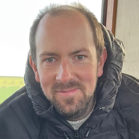 A person wearing a dark puffer jacket, sitting indoors with a green landscape visible through the window.