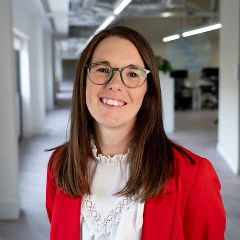 A woman wearing a red jacket and white blouse stands in a modern office space with a bright and inviting atmosphere.