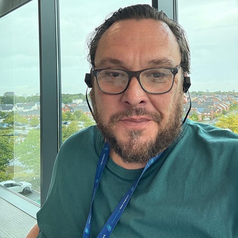 A person wearing headphones and a green t-shirt, sitting by a large window with a view of urban architecture and greenery.