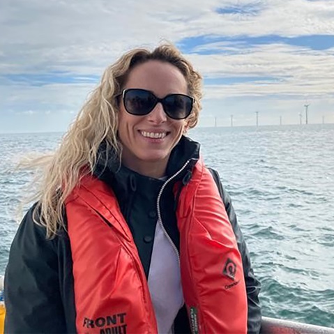 A person wearing a black jacket and a bright orange life jacket stands by the sea with wind turbines visible in the background.