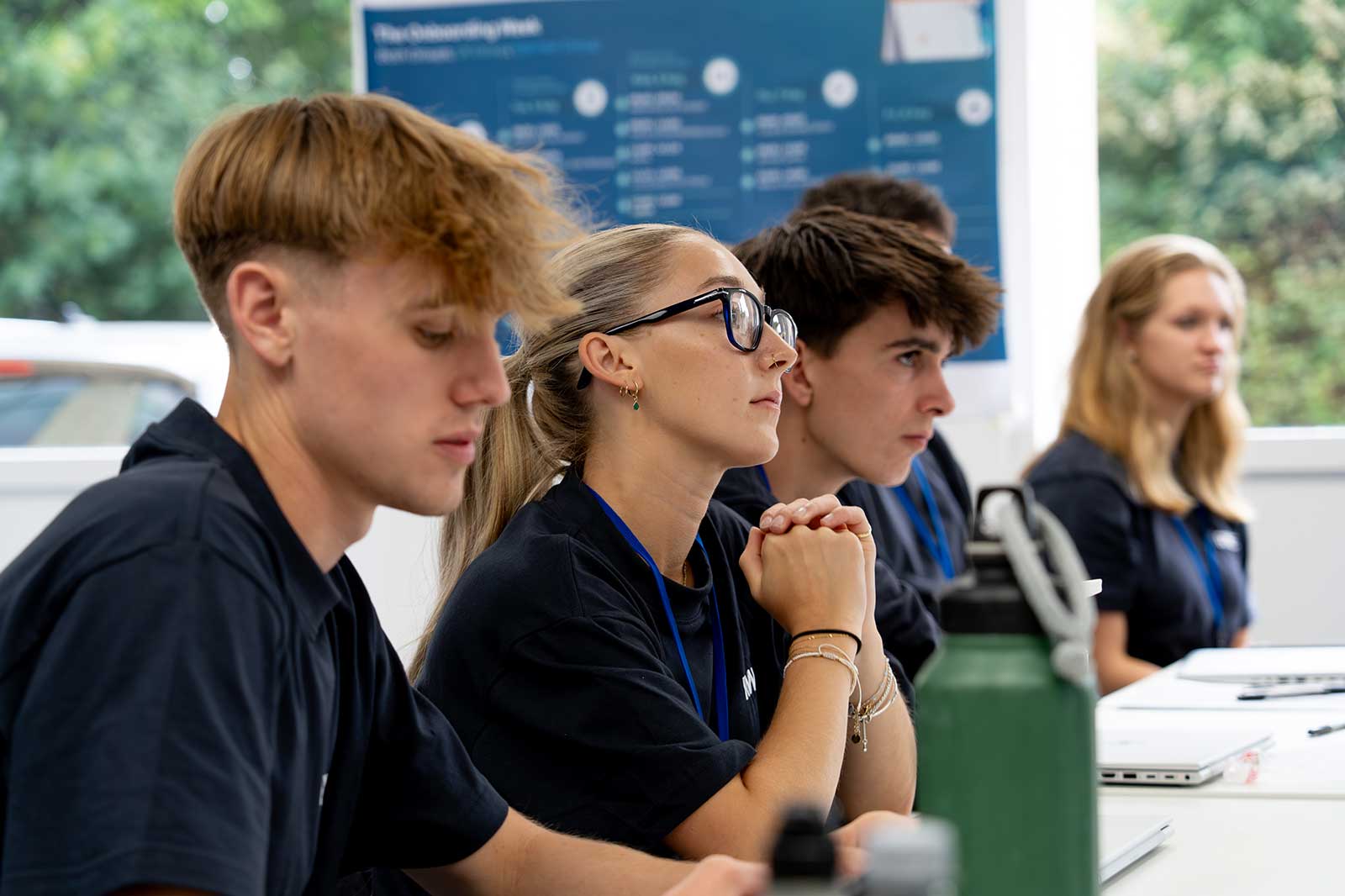 A group of students attentively listening during a session, wearing matching black shirts. A water bottle and laptop are visible.