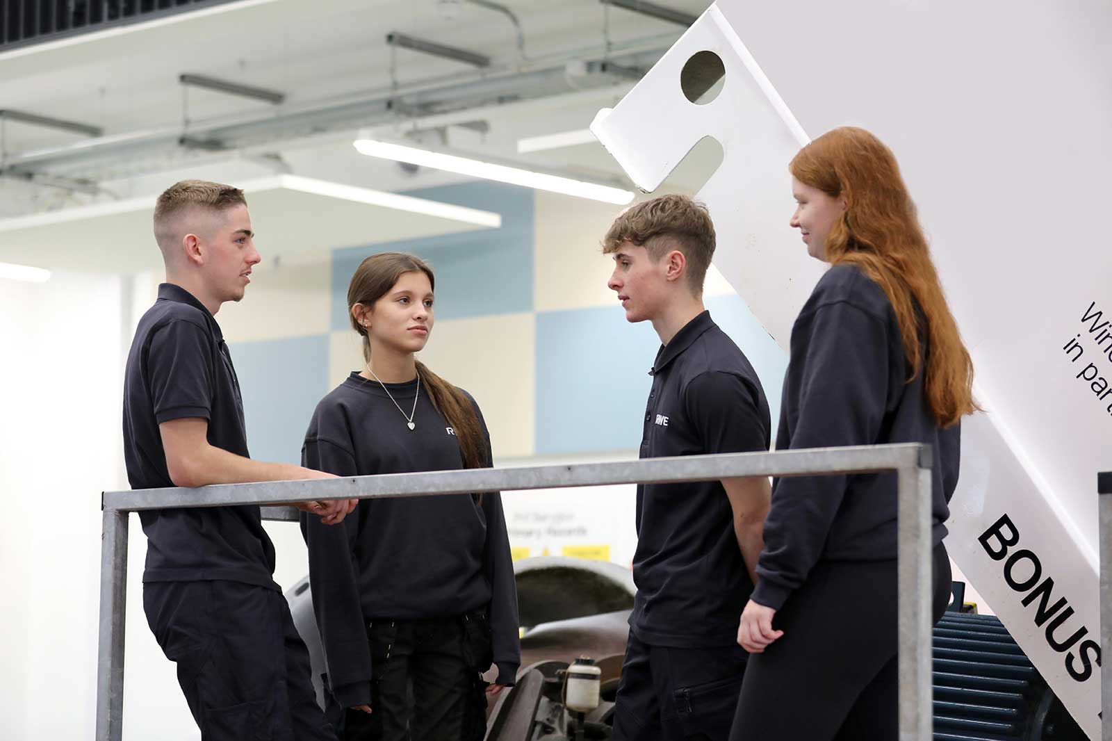 Four individuals in black outfits are engaged in conversation near a car workshop, with an industrial backdrop.