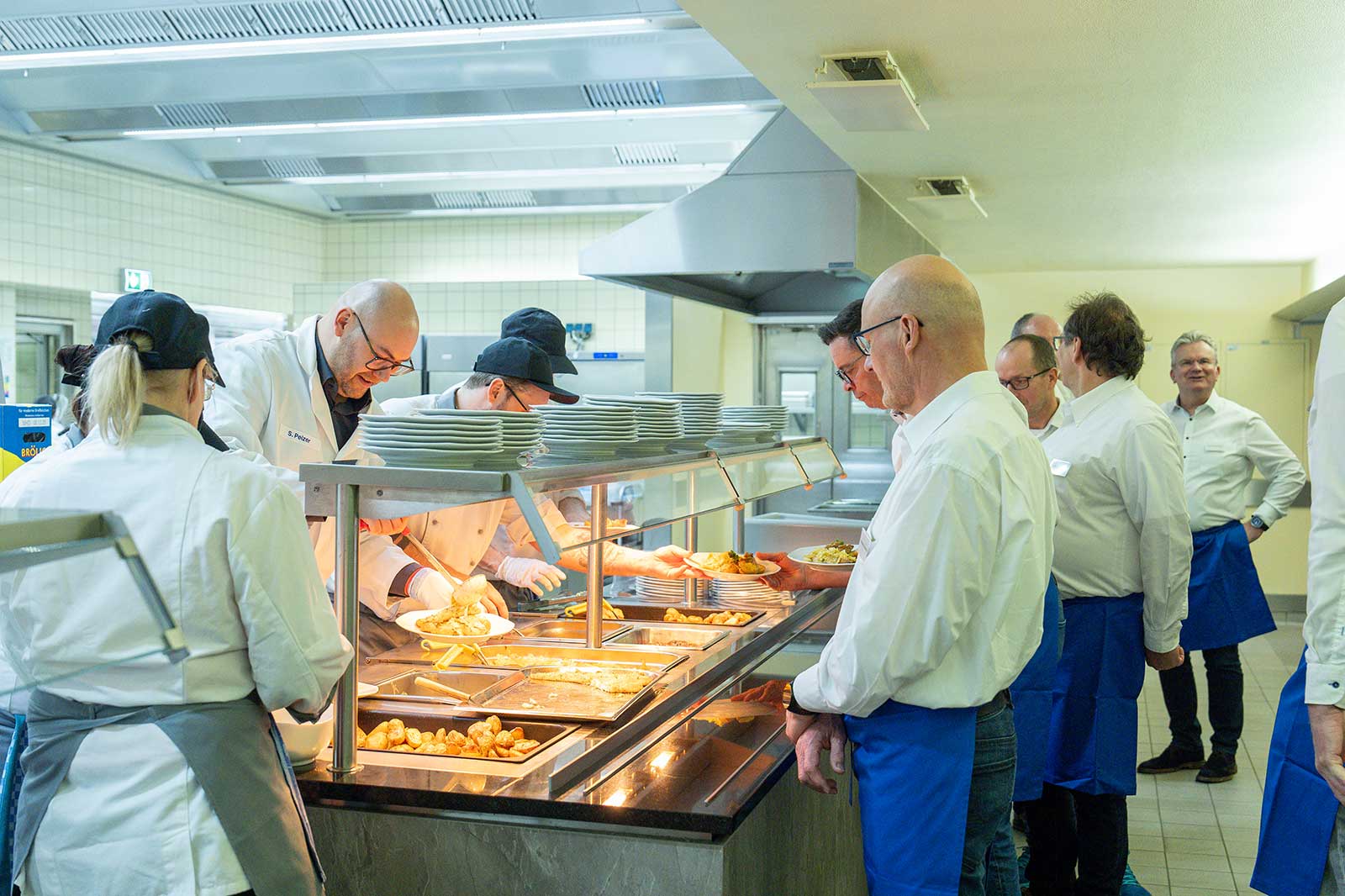 A busy kitchen scene with chefs serving meals at a buffet, while diners await their food in the background.