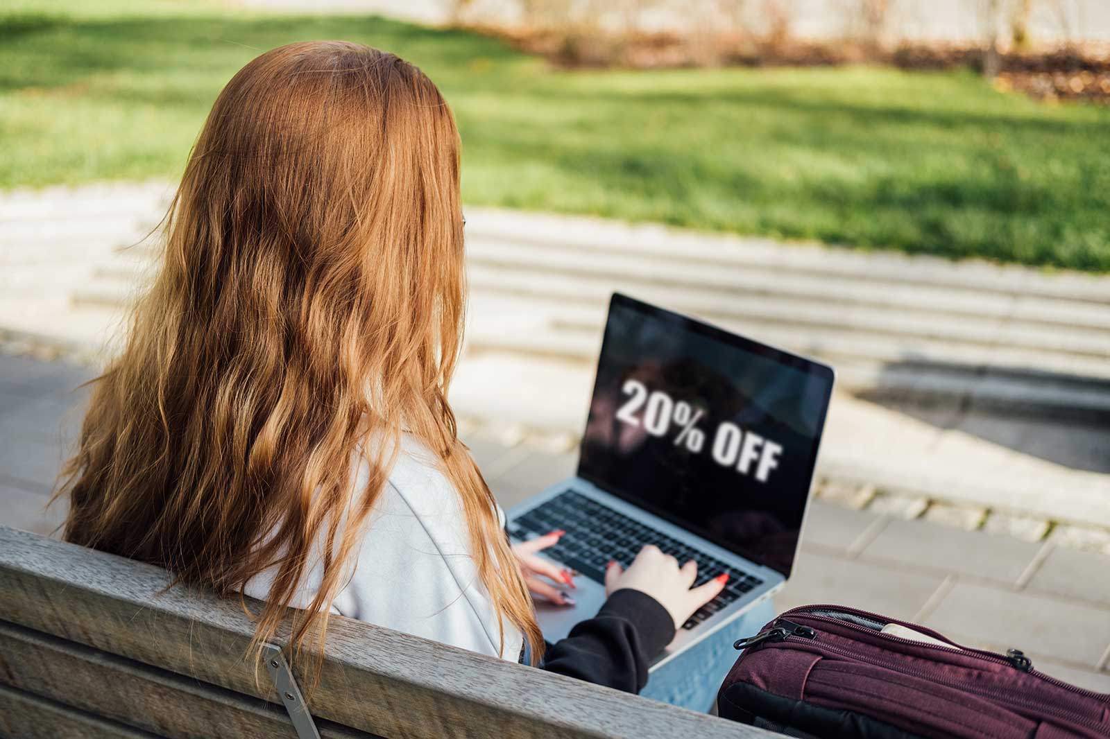 A young woman with wavy, auburn hair sits on a bench, using a laptop displaying '20% OFF' outdoors.