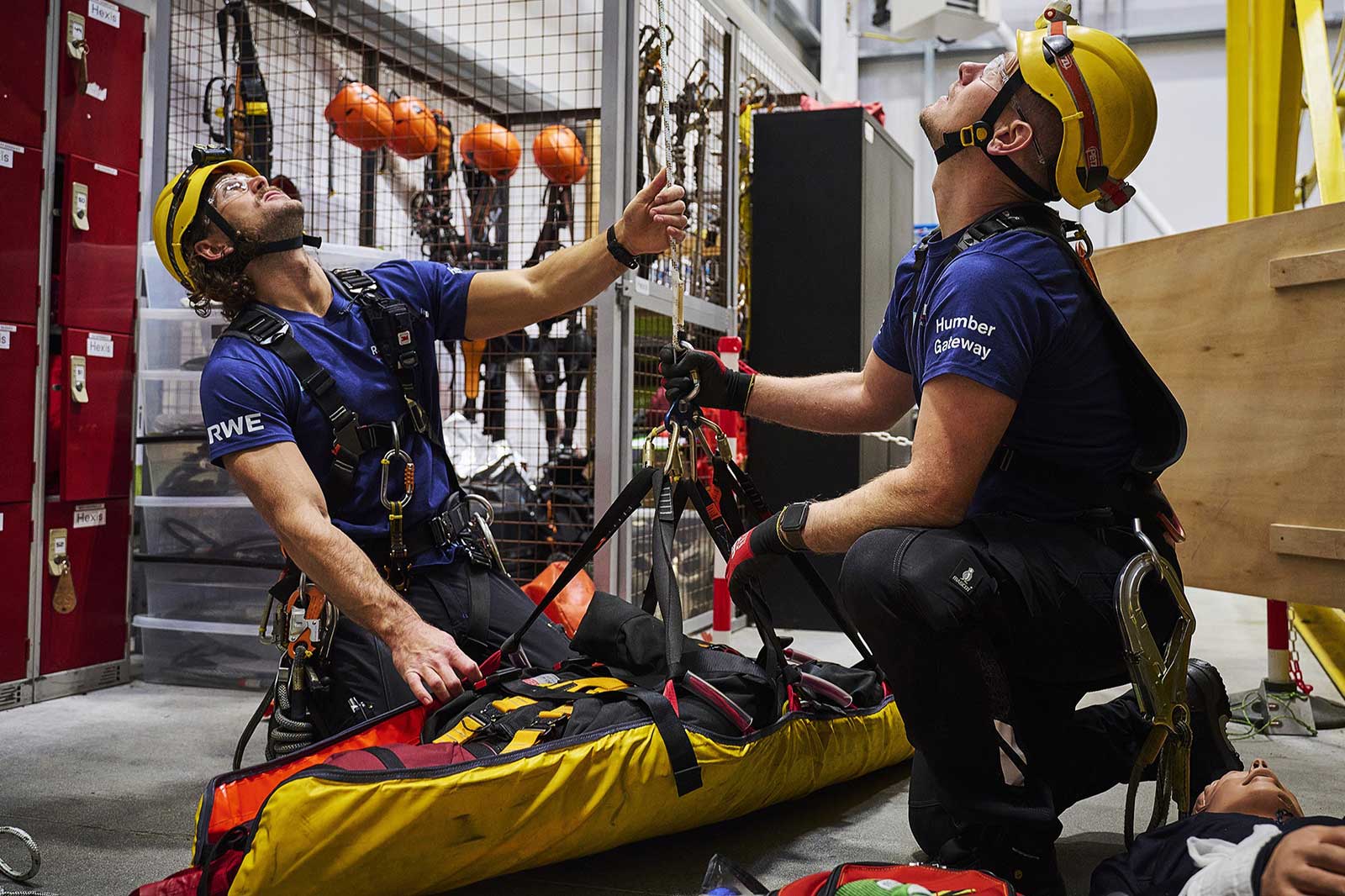 Two rescue workers in helmets prepare equipment in a safety training facility. A rescue stretcher lies on the ground.