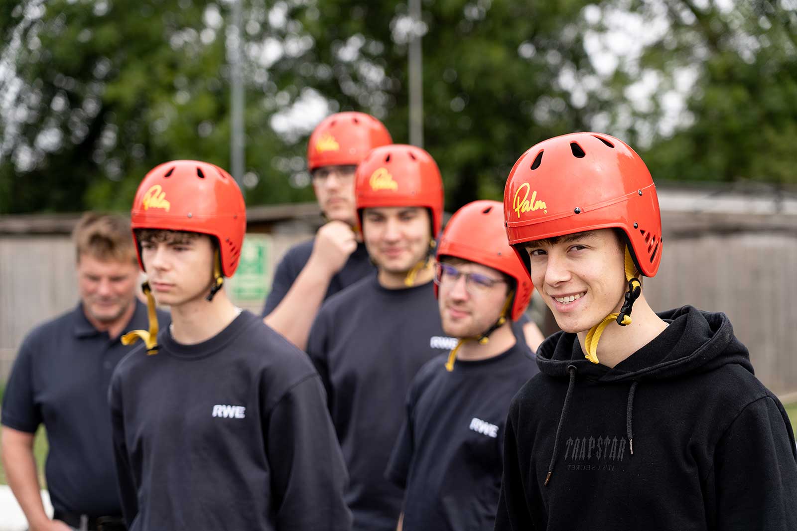 A group of individuals wearing red helmets stands in a line, preparing for an outdoor activity. The background features greenery.