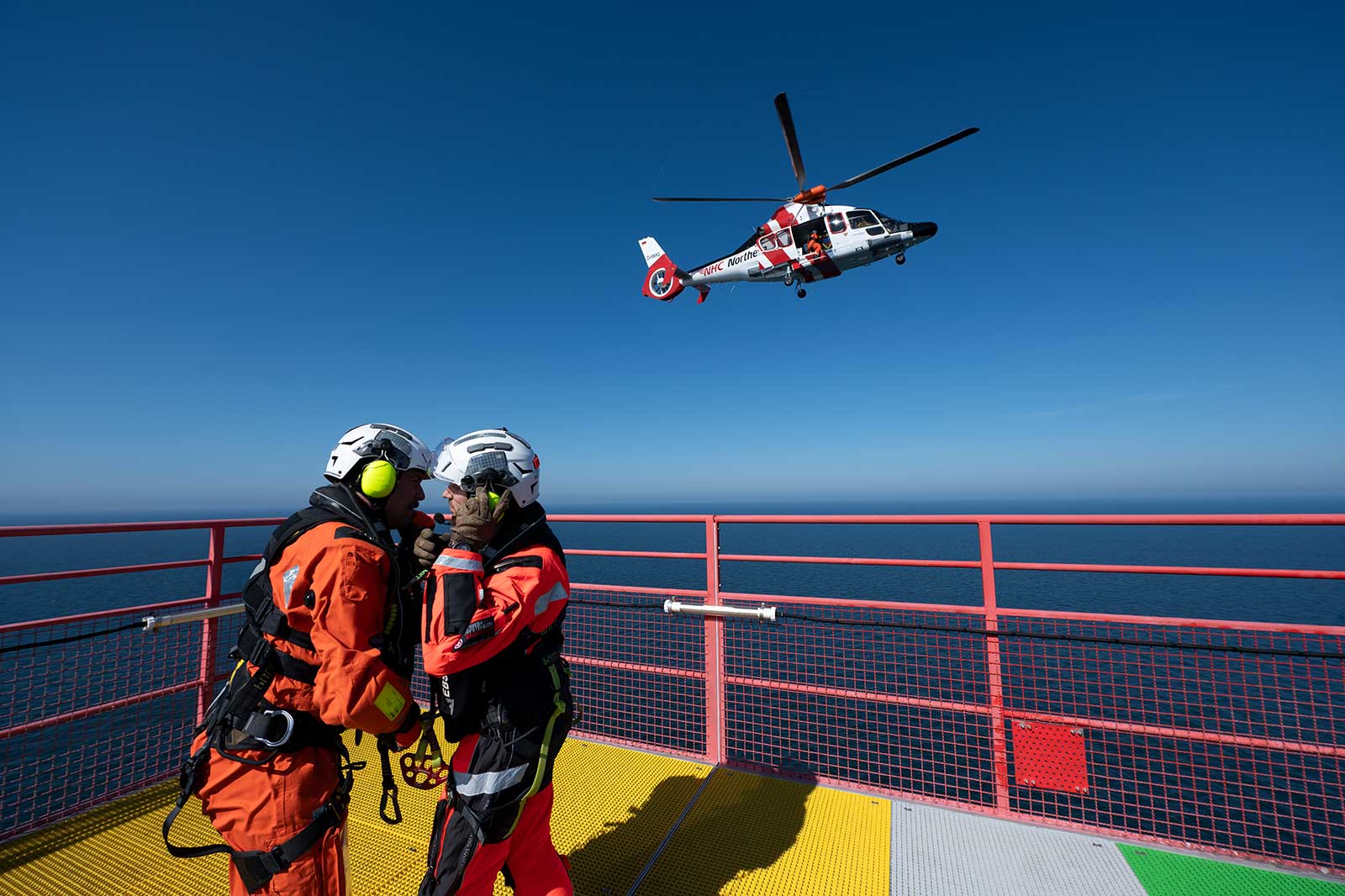 Two rescue workers in orange gear prepare for a helicopter operation against a clear blue sky above the ocean.